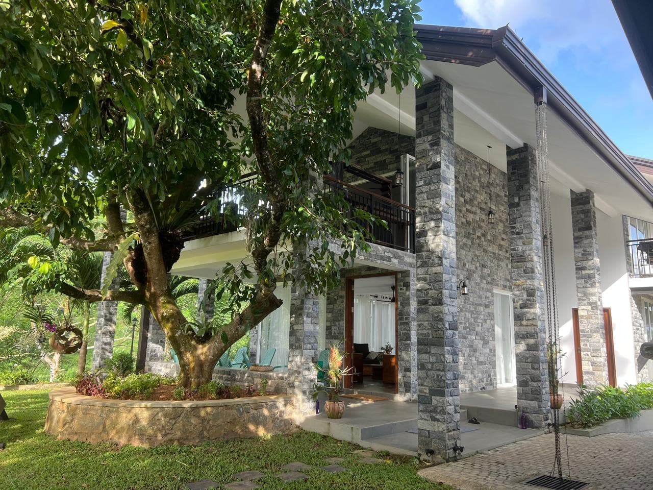 Two-story bungalow in daylight with grey stone pillars, a large shade tree, and a paved walkway